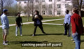 A group of college students standing on a grassy lawn in front of a large white building. One person in the center is holding a volleyball and demonstrating a play, with the subtitle 'catching people off guard.'