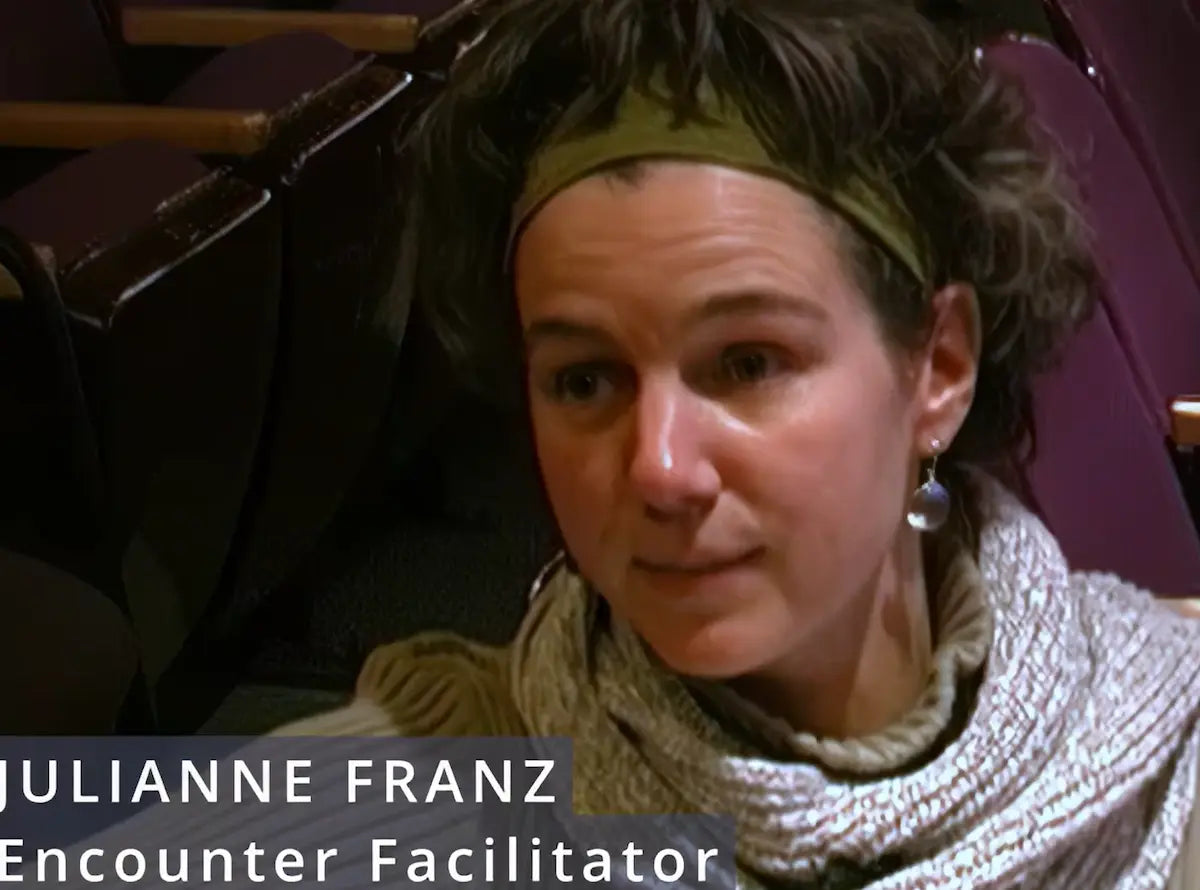 A close-up portrait of Julianne Franz, short curly brown hair, wearing an olive green headband and a chunky knit beige sweater. She is seated in a dark, dimly lit environment with purple seating visible in the background.