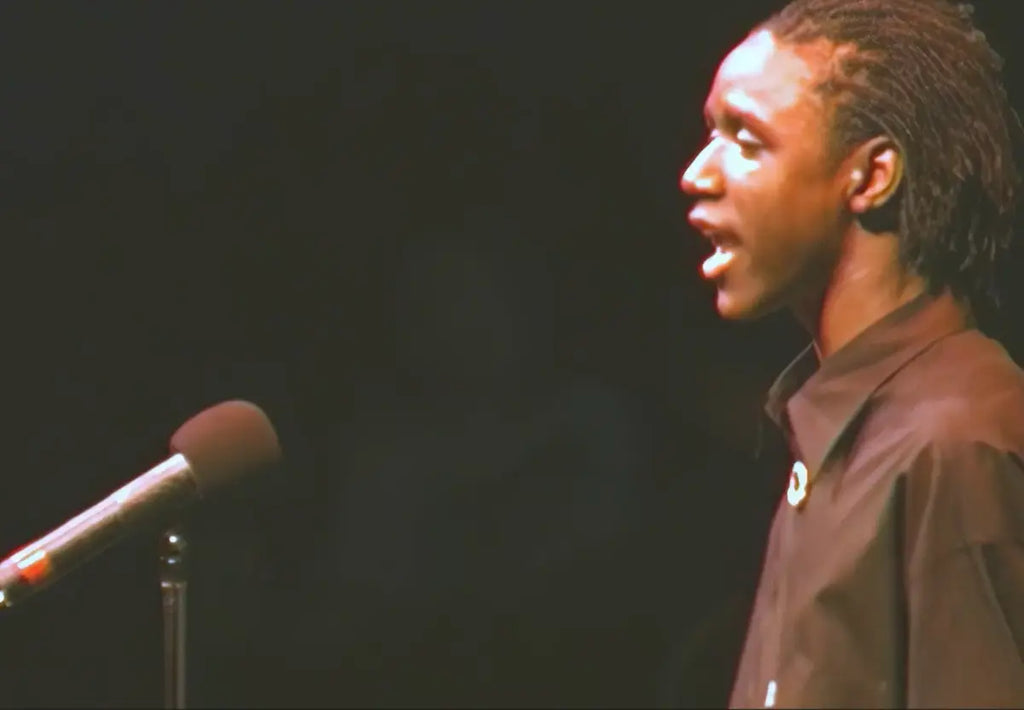 A high school poet with dreadlocks speaking into a microphone, wearing a dark brown collared shirt against a dark background.