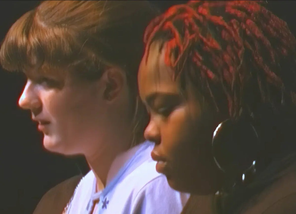 Two young women shown in close profile against a dark background, with one wearing a white top and the other wearing a dark top. The image is tightly cropped and has a warm, low-light tone.