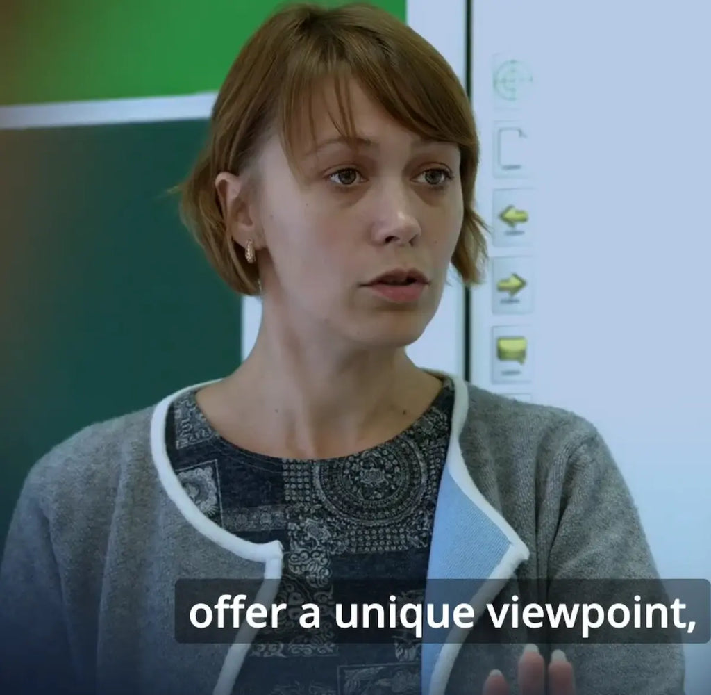 A woman with short auburn hair wearing a gray cardigan, speaking in front of a whiteboard. A subtitle at the bottom reads 'offer a unique viewpoint'.